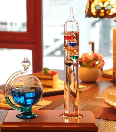 Termómetro de Galileu com esfera barométrica visível, vidro transparente, líquidos coloridos e escala de medição, colocado numa mesa de sala com iluminação quente e decoração de interior.