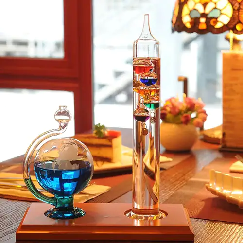 Termómetro de Galileu com esfera barométrica visível, vidro transparente, líquidos coloridos e escala de medição, colocado numa mesa de sala com iluminação quente e decoração de interior.