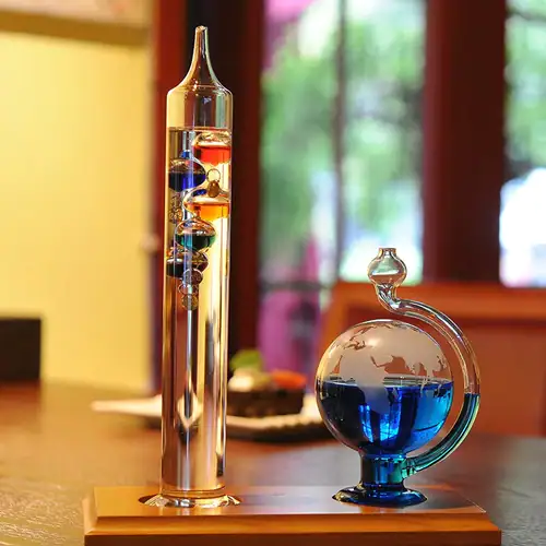 Termómetro de Galileu com esfera barométrica de vidro, contendo líquidos coloridos e esferas internas, colocado numa mesa de madeira com fundo de janela e cortinas verdes.