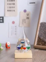 Termómetro de Galileu com gotas de diferentes cores suspensas em um tubo de vidro, apoiado numa base de madeira, com fundo de parede com notas e quadro de café.