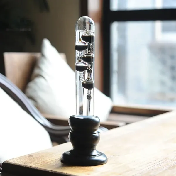 Termómetro de Galileu com esferas de metal suspensas em tubo de vidro, apoiado numa base preta, em ambiente com luz natural e mobiliário de madeira.