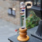 Termómetro de Galileu com esferas de cores diferentes, visíveis através de um tubo de vidro, apoiado numa base de madeira, ao ar livre.