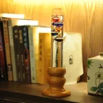 Termómetro de Galileu com tubo de vidro, bolhas coloridas e base de madeira, colocado numa estante com livros e decoração de ciência ao fundo.