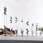 Vários tubos de ensaio de vidro com líquidos de cores diferentes, de diferentes tamanhos, alinhados numa mesa branca, com livros e plantas ao fundo.