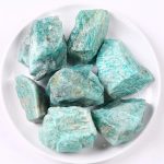 Vários pedaços de amazonite em bruto com tonalidades verdes e azuis, textura irregular e superfícies ásperas, dispostos numa taça branca.