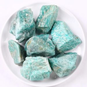 Vários pedaços de amazonite em bruto com tonalidades verdes e azuis, textura irregular e superfícies ásperas, dispostos numa taça branca.