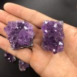Duas amostras de ametista escura em bruto, com cristais violetas e estrutura irregular, mostradas na palma da mão para escala.