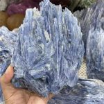 Cianite azul em bruto com cristais alongados e textura fibrosa, exibida na mão para mostrar o tamanho e a estrutura mineral.