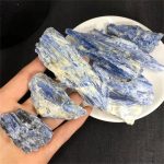 Vários cristais de cianite azul bruta, com diferentes tamanhos e formas, alguns com tonalidades mais claras e outros mais escuros, sobre uma mão e uma tigela branca.