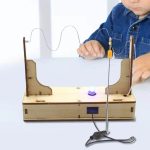 Estudante manipula fios elétricos conectados a uma lâmpada de plasma em experimento educativo, com estrutura de madeira e componentes eletrônicos visíveis.