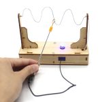 Fio elétrico com lâmpada LED acesa, montagem em base de madeira com componentes eletrônicos, mão segurando o fio, fundo branco.
