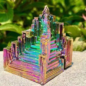 Cristal de Bismuto com estrutura em escadas e cores iridescentes, colocado ao ar livre com vegetação ao fundo.