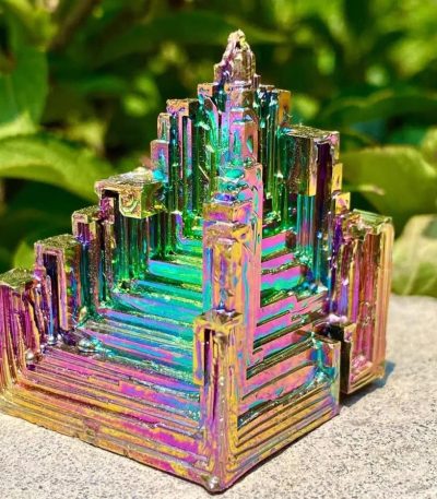 Cristal de Bismuto com estrutura em escadas e cores iridescentes, colocado ao ar livre com vegetação ao fundo.