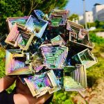 Cristal de bismuto com estrutura em escadas e cores iridescentes, visto ao ar livre com fundo de árvores e edifícios.