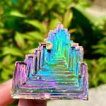 Cristal de Bismuto com estrutura escalonada e cores iridescentes, exibido ao ar livre com fundo de vegetação verde.