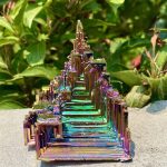 Cristal de bismuto com estrutura em escadaria, refletindo cores iridescentes, colocado sobre uma superfície de pedra com vegetação ao fundo.
