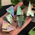Vários cristais de bismuto com estrutura em escadinha iridescente, exibindo cores brilhantes e reflexos, segurados na mão contra fundo de plantas verdes.