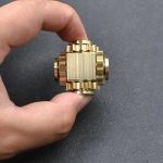 Cubo spinner de mão em metal dourado com detalhes em relevo, visto de cima, com fundo escuro, mostrando o design compacto e detalhes do produto.