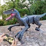 Dinossauro robô com escamas azuis, olhos laranja e boca aberta, controlado por um comando à distância com mãos visíveis na imagem, em ambiente ao ar livre com árvores ao fundo.