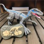 Dinossauro de brinquedo com escamas azuis e cinzentas, olhos laranja, boca aberta mostrando dentes, apoiado numa mesa de madeira, com um modelo de esqueleto de dinossauro ao lado.