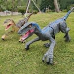 Duas figuras de dinossauros de brinquedo, um T-Rex cinzento e um Velociraptor castanho, colocados em um parque com relva e árvores ao fundo.