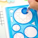 Espirógrafo clássico com engrenagem azul, criando padrões coloridos em papel branco, com uma mão a usar uma caneta para desenhar.