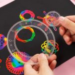 Espirógrafo transparente com várias rodas dentadas, usado para criar desenhos de mandala coloridos, com uma mão segurando uma das rodas.