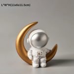Estatueta de astronauta com capacete prateado, apoiado numa lua crescente dourada, com detalhes em branco e dourado, em fundo neutro.