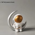 Estatueta de astronauta com capacete dourado, apoiado numa lua prateada em forma de meia-lua, com fundo cinzento neutro.