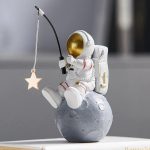 Estatueta de astronauta em pose de pesca, segurando uma vara com uma estrela pendurada, sentado numa esfera que representa a Lua, com detalhes dourados no capacete e mochila.