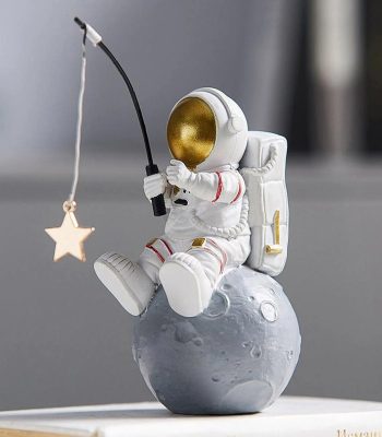 Estatueta de astronauta em pose de pesca, segurando uma vara com uma estrela pendurada, sentado numa esfera que representa a Lua, com detalhes dourados no capacete e mochila.