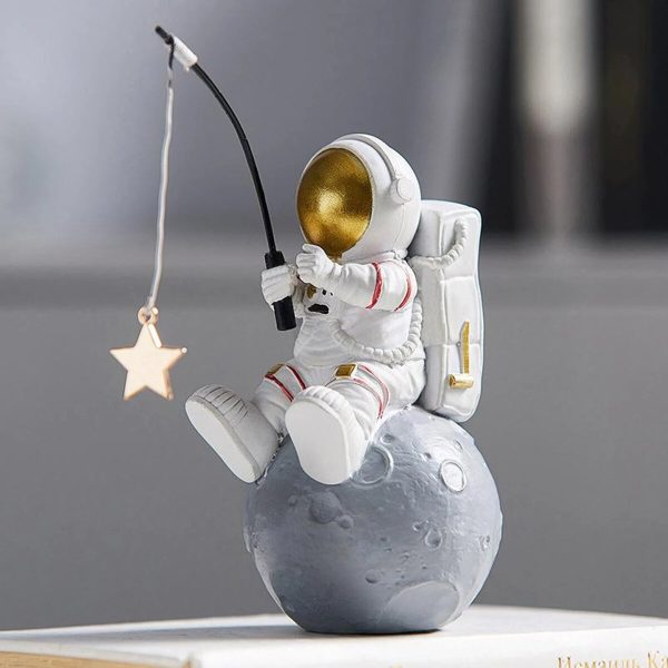 Estatueta de astronauta em pose de pesca, segurando uma vara com uma estrela pendurada, sentado numa esfera que representa a Lua, com detalhes dourados no capacete e mochila.