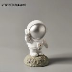 Estatueta de astronauta com capacete prateado, segurando uma caixa, colocada sobre uma base de rocha, com detalhes em branco e prateado.