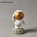 Estatueta de astronauta com capacete dourado, vestuário branco, em pé sobre uma base de rocha, com detalhes de capacete e traje espacial.