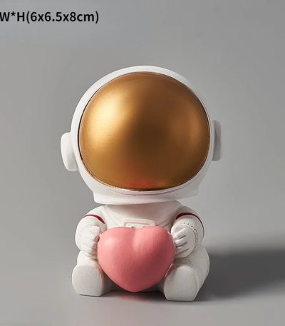 Estatueta de astronauta com capacete dourado e corpo branco, segurando um coração rosa, em posição sentada.