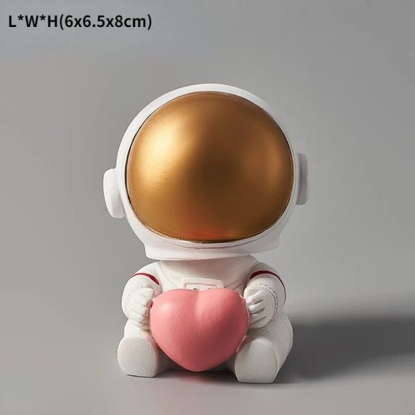 Estatueta de astronauta com capacete dourado e corpo branco, segurando um coração rosa, em posição sentada.