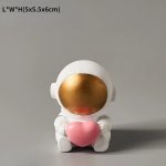 Estatueta de astronauta com capacete branco, rosto dourado e segurando um coração rosa, decorativa e educativa.