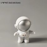 Estatueta de astronauta com capacete prateado e fato branco, com detalhes em cinza, de pé sobre fundo cinzento.