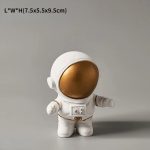 Estatueta de astronauta com capacete dourado, corpo branco e detalhes em dourado, em posição de pé com braços ligeiramente abertos.