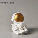 Estatueta de astronauta com capacete dourado e traje branco, sentado numa base, com detalhes em dourado e botões na roupa.