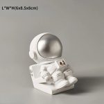 Estatueta de astronauta em miniatura com capacete prateado, sentado numa base branca, com detalhes em dourado no traje.