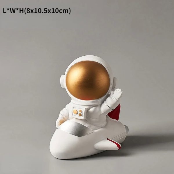 Estatueta de astronauta com capacete dourado, sentado num foguete branco com detalhes vermelhos, em posição de descanso.