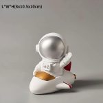 Estatueta de astronauta em traje espacial branco, sentado num foguete decorativo com detalhes dourados e vermelhos, em fundo neutro.