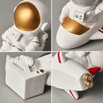 Estatueta de astronauta com capacete dourado, detalhes em ouro na roupa, segurando uma mala branca, com detalhes em vermelho e dourado.