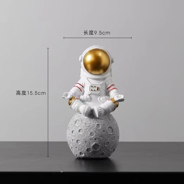 Estatueta de astronauta em posição de meditação, sentado numa base que parece uma lua, com detalhes em branco e dourado na cabeça e roupas.