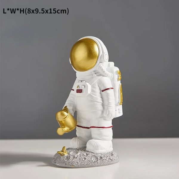 Estatueta de astronauta com capacete dourado, roupa branca com detalhes vermelhos, segurando uma ferramenta dourada, em base de rocha com pequena planta.