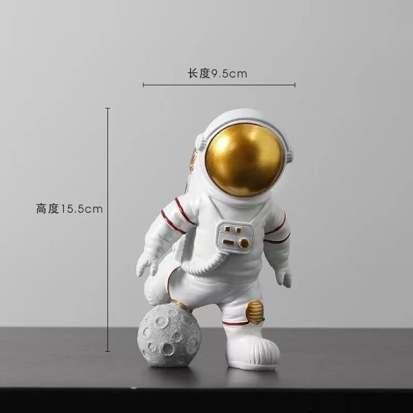 Estatueta de astronauta com capacete dourado, apoiado numa bola que parece a Lua, com detalhes em branco e vermelho, altura de 15,5cm e comprimento de 9,5cm.