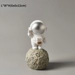 Estatueta de astronauta em traje espacial branco, de pé numa rocha lunar, com capacete prateado e detalhes em dourado na mochila.