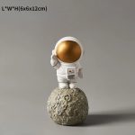 Estatueta de astronauta com capacete dourado, vestuário branco, em pé sobre uma réplica da Lua com textura rochosa, fundo cinzento neutro.