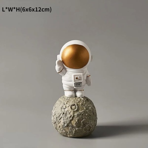 Estatueta de astronauta com capacete dourado, vestuário branco, em pé sobre uma réplica da Lua com textura rochosa, fundo cinzento neutro.